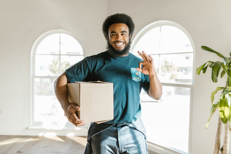 A smiling man with dark curly hair and a beard holds a cardboard moving box in his left hand while making an 'OK' gesture with his right hand indoors, near large arched windows allowing natural light to fill the room. The background shows a bright, modern interior with white walls, a wooden floor, and a potted plant with large green leaves. The scene suggests a home relocation process, with the individual preparing for or having completed packing, aligned with house removals services such as those offered by Man with Van Croydon, as part of furniture transport and moving logistics inside a residential property.