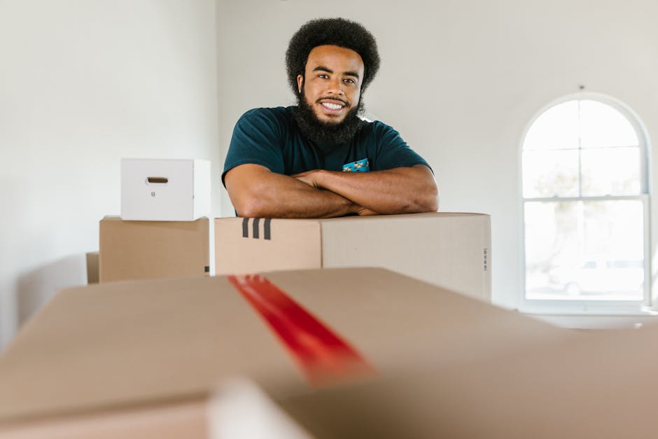 A smiling man with a beard and curly hair standing behind several packed cardboard boxes inside a well-lit room with a large arched window. The boxes vary in size and are sealed with brown packing tape; some contain plastic wrapping and packing materials suitable for a home relocation. The man has his arms crossed on top of a medium-sized box and appears ready for moving or furniture transport as part of a professional removals service. In the background, the indoors environment has light-colored walls, and the natural daylight from the window illuminates the space, indicating a careful packing and loading process typical of home moving activities at an address such as Addisoncombe, supporting the services offered by Man with Van Croydon.