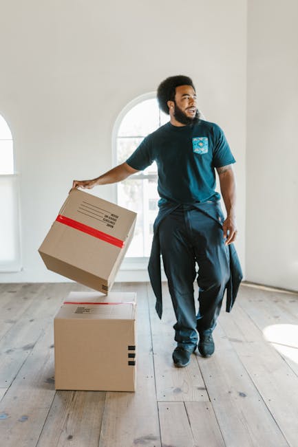 A smiling man with dark curly hair and a beard holds a cardboard moving box in his left hand while making an 'OK' gesture with his right hand indoors, near large arched windows allowing natural light to fill the room. The background shows a bright, modern interior with white walls, a wooden floor, and a potted plant with large green leaves. The scene suggests a home relocation process, with the individual preparing for or having completed packing, aligned with house removals services such as those offered by Man with Van Croydon, as part of furniture transport and moving logistics inside a residential property.