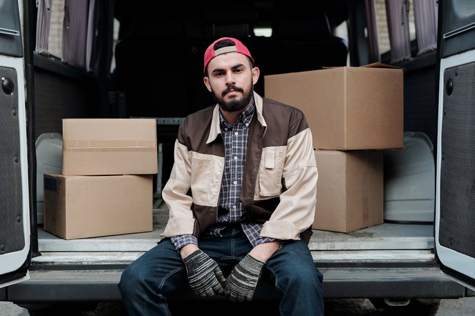 A smiling young male mover with long hair, wearing a black bandana and dark clothing, is sitting in the driver's seat of a white van marked with 'MOVING COMPANY LOCAL & LONG DISTANCE'. The van is park