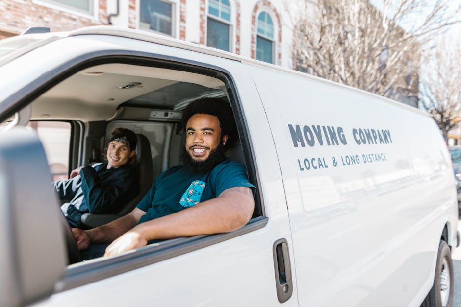 Inside a white moving van labeled 'MOVING COMPANY LOCAL & LONG DISTANCE', two male movers are visible through the open front passenger window. The man in the driver's seat is smiling, has a beard, and is wearing a blue t-shirt with a colourful logo. The passenger, sitting in the front seat, is wearing a dark jacket and a headband, and is also smiling. The van is parked on a street with residential buildings and leafless trees in the background, suggesting a daytime home relocation process. The vehicle is equipped with visible interior features, and the movers appear engaged in preparations for furniture transport and packing and moving tasks, supporting professional house removal services like those offered by Man with Van Croydon.