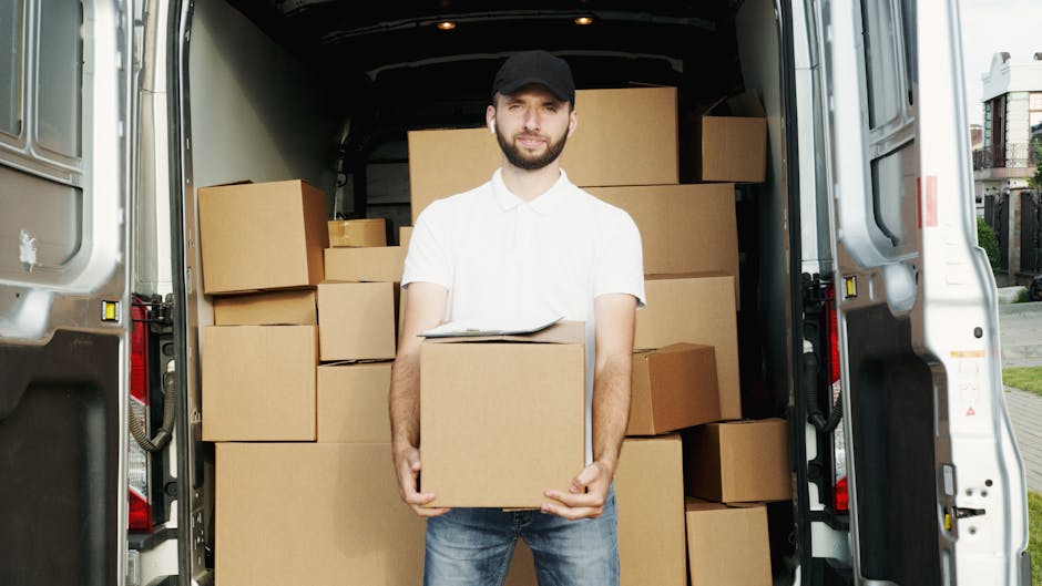 A man wearing a white shirt, dark cap, and jeans is standing at the rear of a large white van, holding a cardboard box during a house removal. The van's interior is filled with stacked cardboard boxes of various sizes, some wrapped in plastic or packing materials, ready for transport. The sequence appears to be part of a furniture transport or home relocation process, with the man positioned on the pavement outside a residential property. The background shows part of a street with houses and greenery, indicating an outdoor loading and unloading environment. The image reflects a professional removals service, exemplifying packing, loading, and moving logistics handled by a company like Man with Van Croydon, highlighting efficient and organized furniture transport.