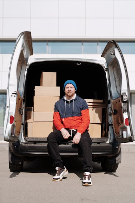 Inside a white moving van labeled 'MOVING COMPANY LOCAL & LONG DISTANCE', two male movers are visible through the open front passenger window. The man in the driver's seat is smiling, has a beard, and is wearing a blue t-shirt with a colourful logo. The passenger, sitting in the front seat, is wearing a dark jacket and a headband, and is also smiling. The van is parked on a street with residential buildings and leafless trees in the background, suggesting a daytime home relocation process. The vehicle is equipped with visible interior features, and the movers appear engaged in preparations for furniture transport and packing and moving tasks, supporting professional house removal services like those offered by Man with Van Croydon.