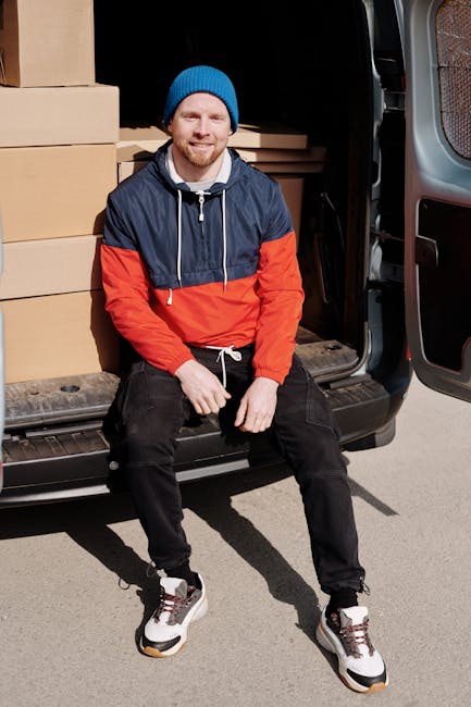 A man wearing a blue beanie, a navy and red waterproof jacket, black pants, and white sneakers with black and red details, sitting on the bumper of a black van with an open sliding door. Inside the van, several large cardboard boxes are visible, some wrapped in plastic or packing paper, arranged neatly for a home relocation. The man is positioned on the pavement, appearing relaxed and smiling, with packaging materials and boxes prepared for furniture transport and moving logistics. The scene reflects the loading process during a professional removals service by Man with Van Croydon, capturing the typical elements of packing, loading furniture, and moving supplies in a residential neighbourhood near East Croydon station.