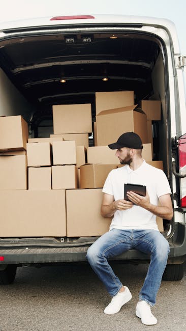 A man wearing a white t-shirt, blue jeans, white sneakers, and a dark cap is sitting on the edge of an open van, which is loaded with multiple cardboard boxes of varying sizes, some of which are sealed with packing tape. The boxes are arranged inside the van, with some stacked on top of each other, indicating a home relocation or furniture transport process. The van's interior reveals a dark, empty cargo space, while the exterior environment appears to be outdoors with natural lighting. The man is holding a tablet or device, possibly reviewing moving details or instructions, suggesting he is involved in the packing and moving process. This scene illustrates the typical loading and logistics associated with house removals and professional moving services, as handled by companies like Man with Van Croydon, during furniture transport and household packing activities.