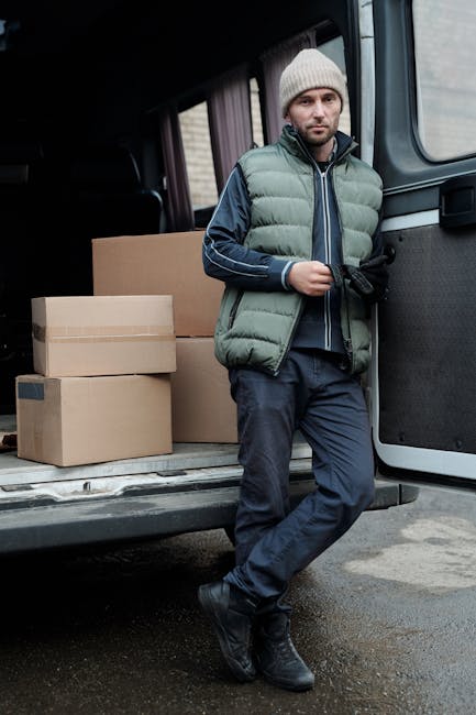A man wearing a blue beanie, a navy and red waterproof jacket, black pants, and white sneakers with black and red details, sitting on the bumper of a black van with an open sliding door. Inside the van, several large cardboard boxes are visible, some wrapped in plastic or packing paper, arranged neatly for a home relocation. The man is positioned on the pavement, appearing relaxed and smiling, with packaging materials and boxes prepared for furniture transport and moving logistics. The scene reflects the loading process during a professional removals service by Man with Van Croydon, capturing the typical elements of packing, loading furniture, and moving supplies in a residential neighbourhood near East Croydon station.