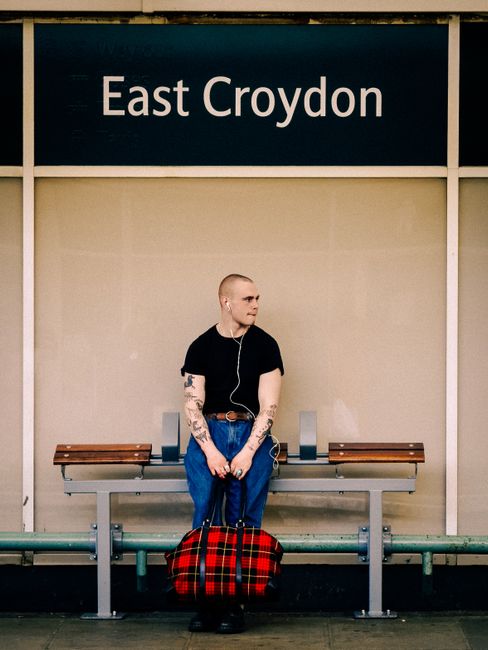 A young man with a shaved head, wearing a black t-shirt, blue jeans, and white earphones, is sitting on a grey metal bench at East Croydon train station platform. He has visible tattoos on both arms a